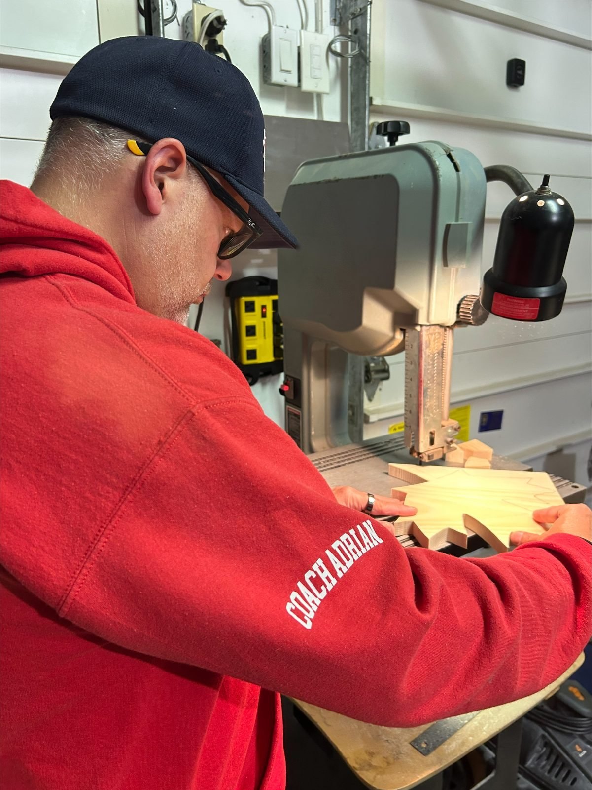 Adrian, wearing a red hoodie and cap, cutting a pine maple leaf by hand on a band saw